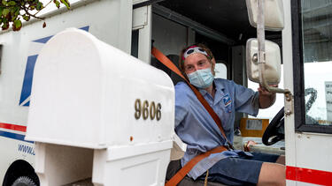 A USPS worker wearing a mask puts envelopes in a mailbox while driving past 