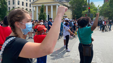 A White protester standing behind a Black protester, both with fists raised.