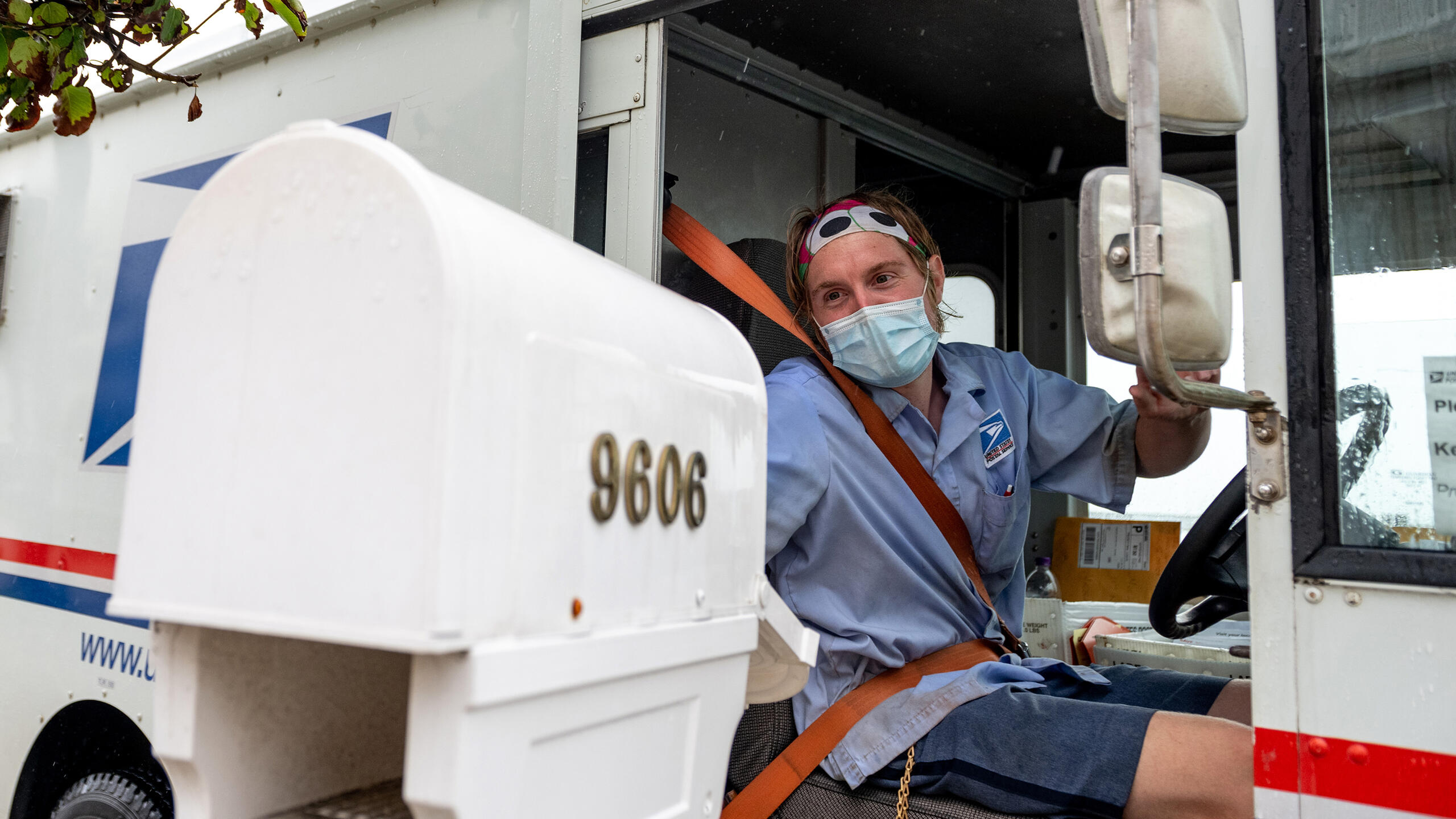 A USPS worker wearing a mask puts envelopes in a mailbox while driving past