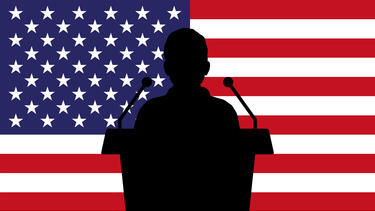 A silhouette of a speaker at a podium in front of an American flag
