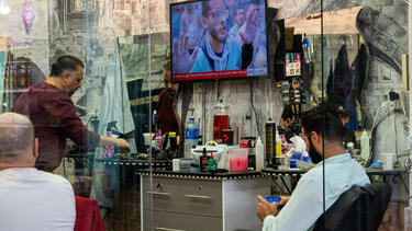 A man looking at his phone in front of a television showing scenes from the Israel-Hamas war.