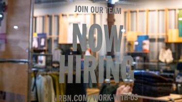 A "Now Hiring" sign in the window of a mall clothing store