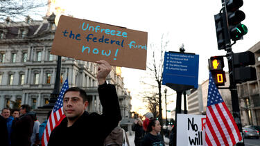 A protester holding a sign reading "unfreeze the federal funds now."