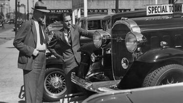 A black and white photo of a car salesman talking to a customer about a mid-20th-century car