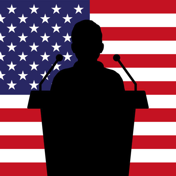 A silhouette of a speaker at a podium in front of an American flag