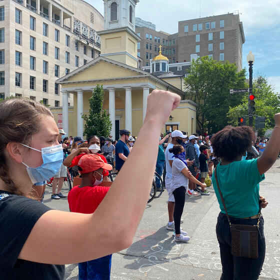 A White protester standing behind a Black protester, both with fists raised.