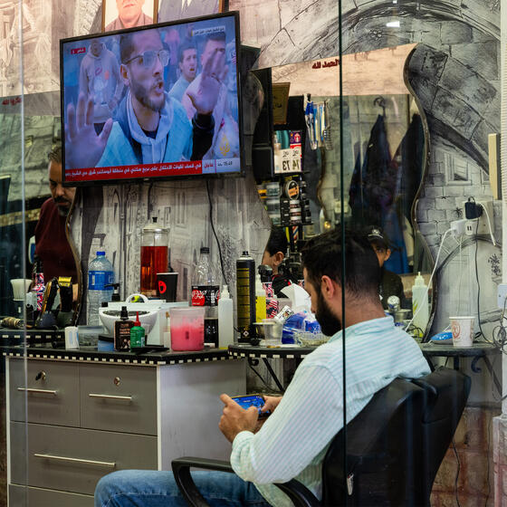 A man looking at his phone in front of a television showing scenes from the Israel-Hamas war.