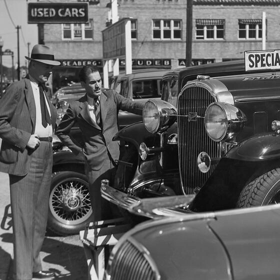 A black and white photo of a car salesman talking to a customer about a mid-20th-century car