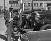 A black and white photo of a car salesman talking to a customer about a mid-20th-century car